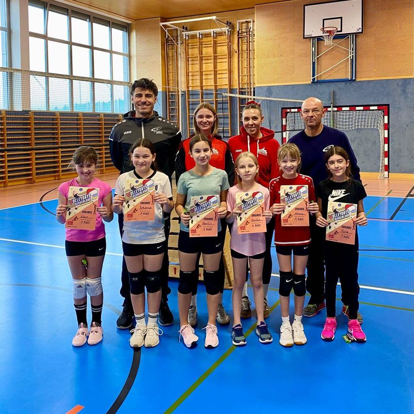 Eine Gruppe junger Mädchen mit Urkunden posiert für ein Foto in einer Turnhalle mit drei Erwachsenen.