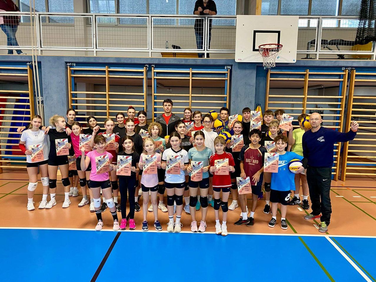 Eine Gruppe junger Athleten und Trainer steht auf einem Spielfeld, hält Urkunden in den Händen und lächelt für ein Foto. Im Hintergrund sind ein Basketballkorb und Holzstangen zu sehen.