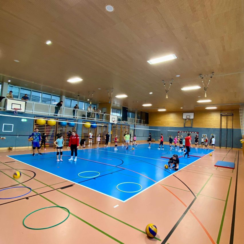 Eine Indoor-Sportanlage mit einem blauen Spielfeld, verschiedenen farbigen Bällen und mehreren spielenden Kindern. Das Spielfeld hat Markierungen und eine Person kniet. Die Decke hat Lampen und ein Geländer.