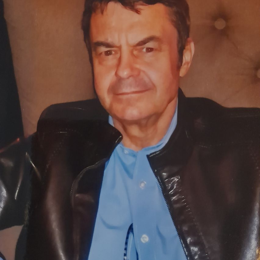 Ein Mann mit kurzen braunen Haaren trägt ein blaues Hemd und eine schwarze Lederjacke. Er sitzt auf einem braunen Ledersofa.