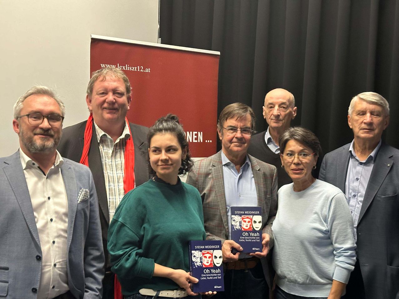 Gruppenfoto von Personen bei einer Veranstaltung, die Bücher halten, mit einem roten Banner hinter ihnen. Das Banner hat weißen Text, der 'www.lexiszt12.at' lautet.