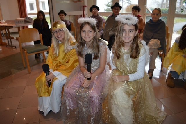 Drei Mädchen in Kostümen, eines mit einem Mikrofon, sitzen vor einer Gruppe von Menschen. Sie führen wahrscheinlich ein Theaterstück auf.