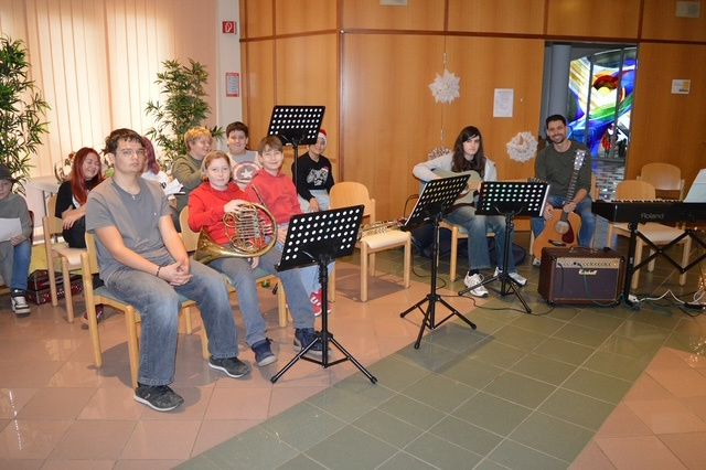 Eine Gruppe von Menschen sitzt in einem Raum, einige spielen Instrumente und andere beobachten. Stühle und Notenständer sind vorhanden.