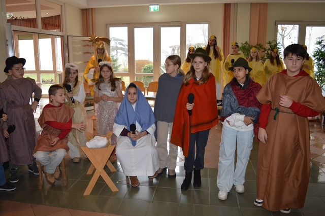 Kinder in Kostümen versammeln sich in einem Raum mit einer Krippenszene, halten Mikrofone und posieren für ein Gruppenfoto.