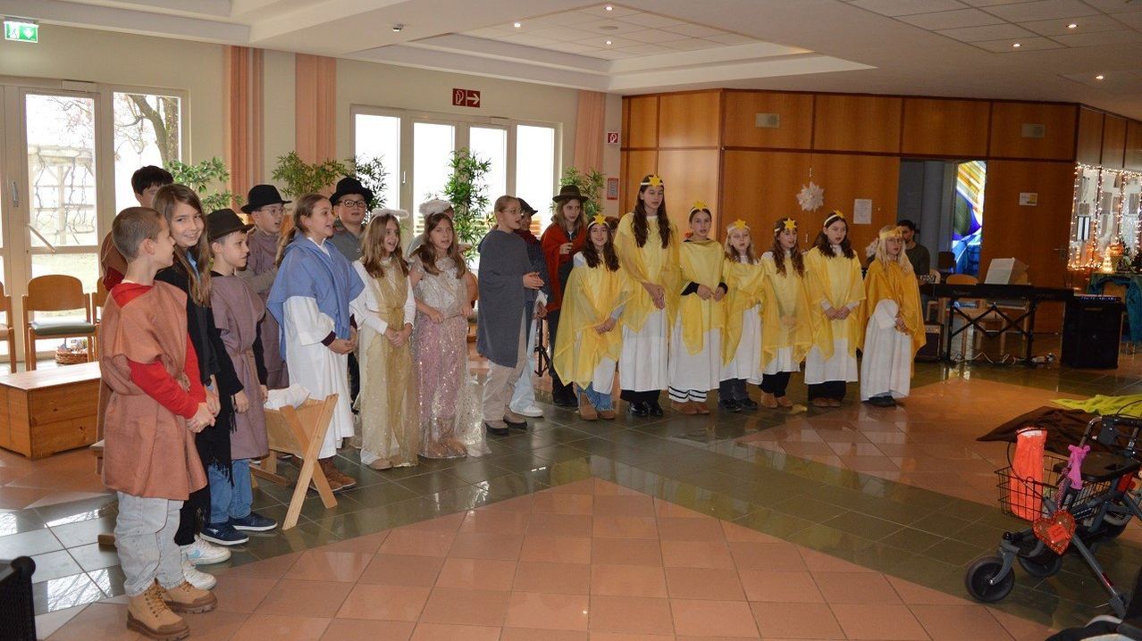Eine Gruppe von Kindern in Kostümen mit Sternen auf den Köpfen steht in einer Reihe in einem Raum mit gefliesten Böden. Dahinter stehen Erwachsene, von denen einige ebenfalls Kostüme tragen. Im Hintergrund befinden sich Pflanzen und eine Holzbank.