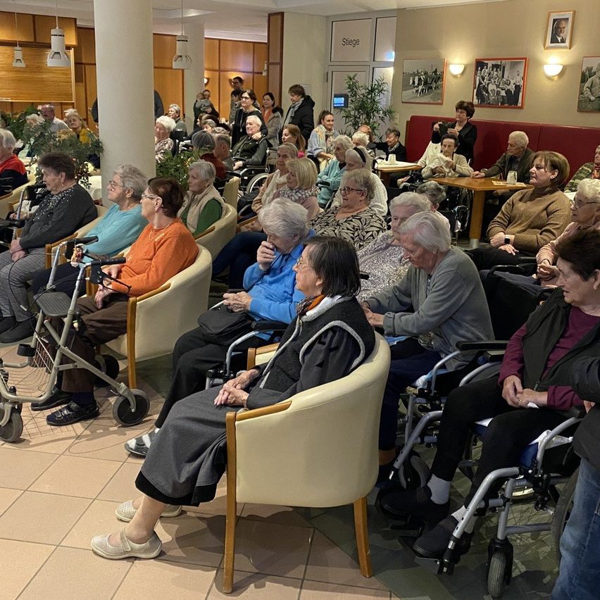 Eine Gruppe älterer Menschen sitzt in Stühlen in einem Raum. Einige sind im Rollstuhl. Sie unterhalten sich. Es gibt eine Säule und eine Pflanze im Raum.
