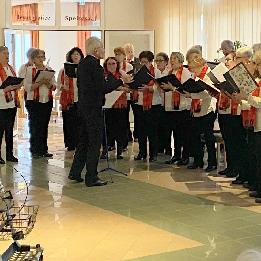 Eine Gruppe älterer Frauen und Männer singt zusammen, hält Musikbücher in den Händen und steht in einem Raum. Ein Mann im Anzug steht vor ihnen.