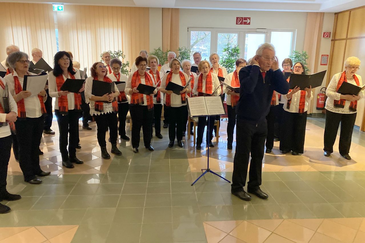 Ein Chor singt in einem Raum mit gefliestem Boden, trägt Schals und hält Musikbücher. Der Dirigent steht vor ihnen und hält einen Notenständer.