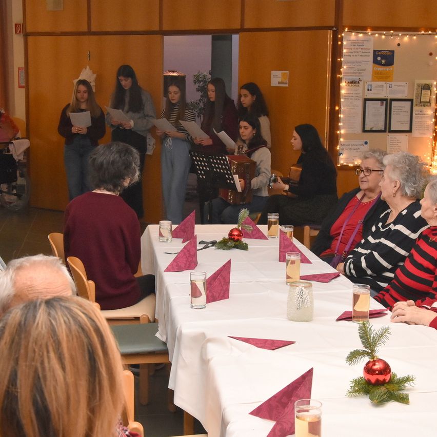 Eine Gruppe von Frauen und Mädchen singt vor einem sitzenden Publikum, möglicherweise in einem Altersheim. Das Publikum besteht aus älteren Menschen und vor ihnen steht ein Tisch mit Getränken. Der Raum ist mit Weihnachtslichtern und Papieren an der Wand geschmückt.