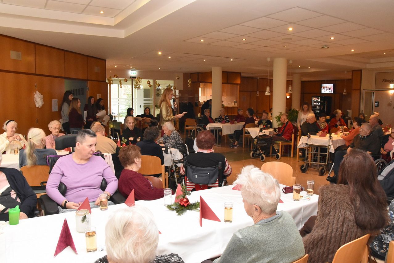 Eine Gruppe älterer Frauen versammelt sich in einem Speisesaal, einige sitzen und andere stehen. Eine Frau steht an einem Tisch mit Mikrofon und spricht die Gruppe an. Der Raum ist mit Weihnachtsdekoration geschmückt.