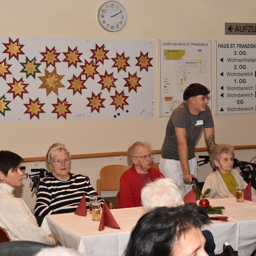 Eine Gruppe älterer Frauen sitzt in einem Speisesaal um einen Tisch, ein Mann steht hinter ihnen. Eine Uhr und ein Grundriss hängen an der Wand.