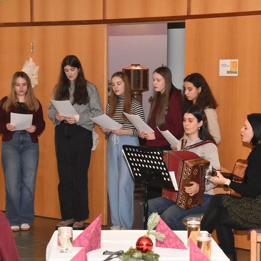 Eine Gruppe junger Frauen steht und hält Papiere in einem Raum. Eine Frau sitzt auf einem Stuhl und spielt Akkordeon. Vor ihnen steht ein Tisch mit einem Weinglas und einer roten Kugel.