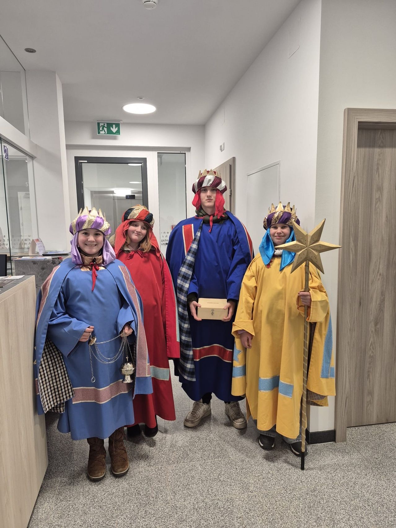 Fünf Personen in Kostümen, wahrscheinlich für ein Weihnachtsspiel, stehen in einem Flur. Drei Frauen und zwei Männer. Der Mann in der Mitte hält eine Schachtel.