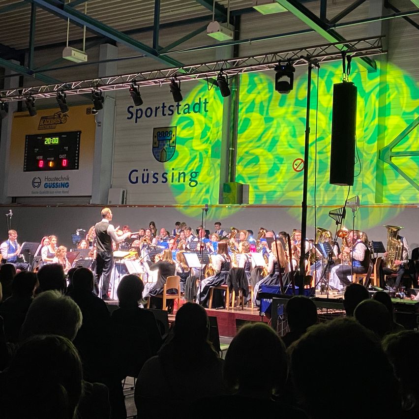 Ein Orchester spielt auf der Bühne in der Sportstadt Gussing, vor einem sitzenden Publikum. Die Bühne ist mit einer grünen Lichtprojektion beleuchtet.