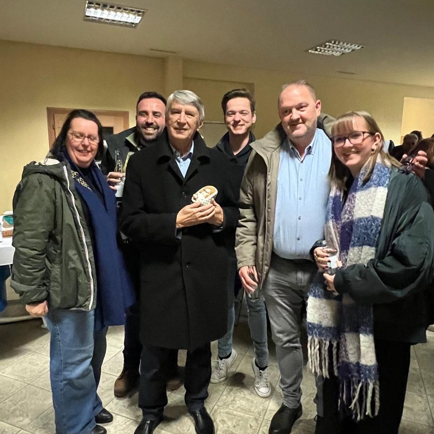 Eine Gruppe von Menschen in einem Raum, lächelnd und für ein Foto posierend. Der Mann in der Mitte hält ein Sandwich, während andere Getränke haben. Sie tragen Winterkleidung.