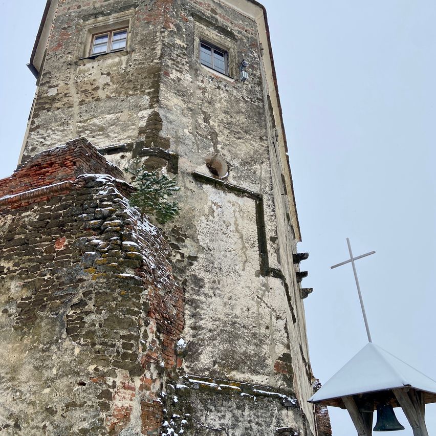 Ein alter Turm mit zwei Fenstern, verwitterten Mauern und einem Glockenturm. Schnee liegt auf dem Ziegeldach und ein Kreuz schmückt den Glockenturm.