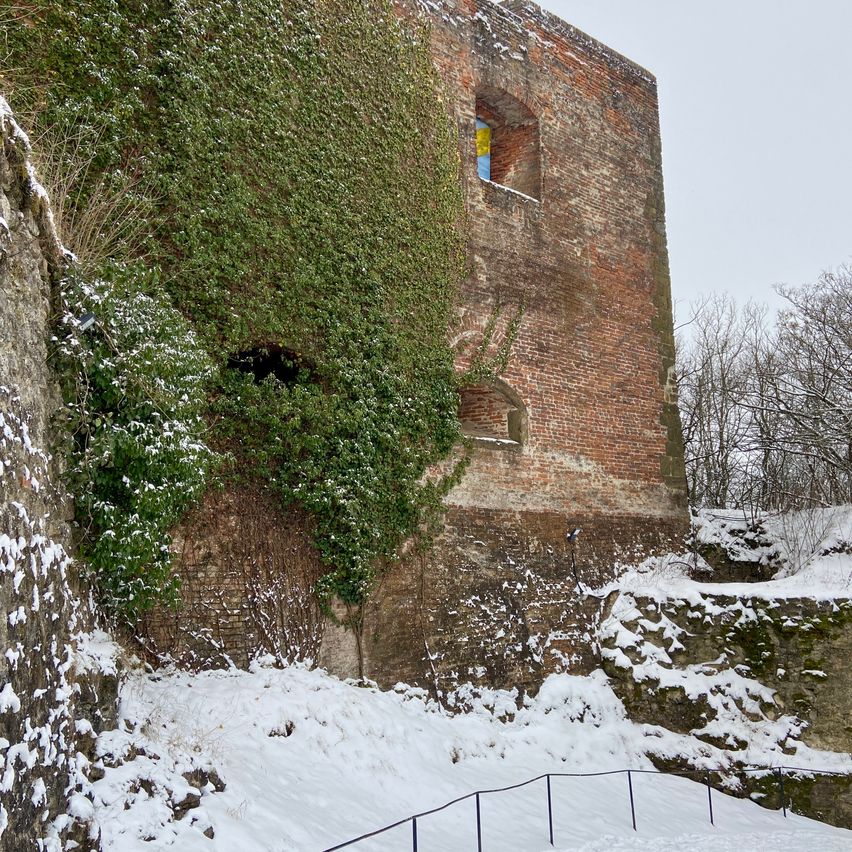 Ein alter Backsteinturm mit von Efeu bewachsenen Wänden steht in einer verschneiten Landschaft. Der Turm hat ein kleines Fenster und ist teilweise von Schnee bedeckt. In der Nähe befindet sich ein Metallzaun.