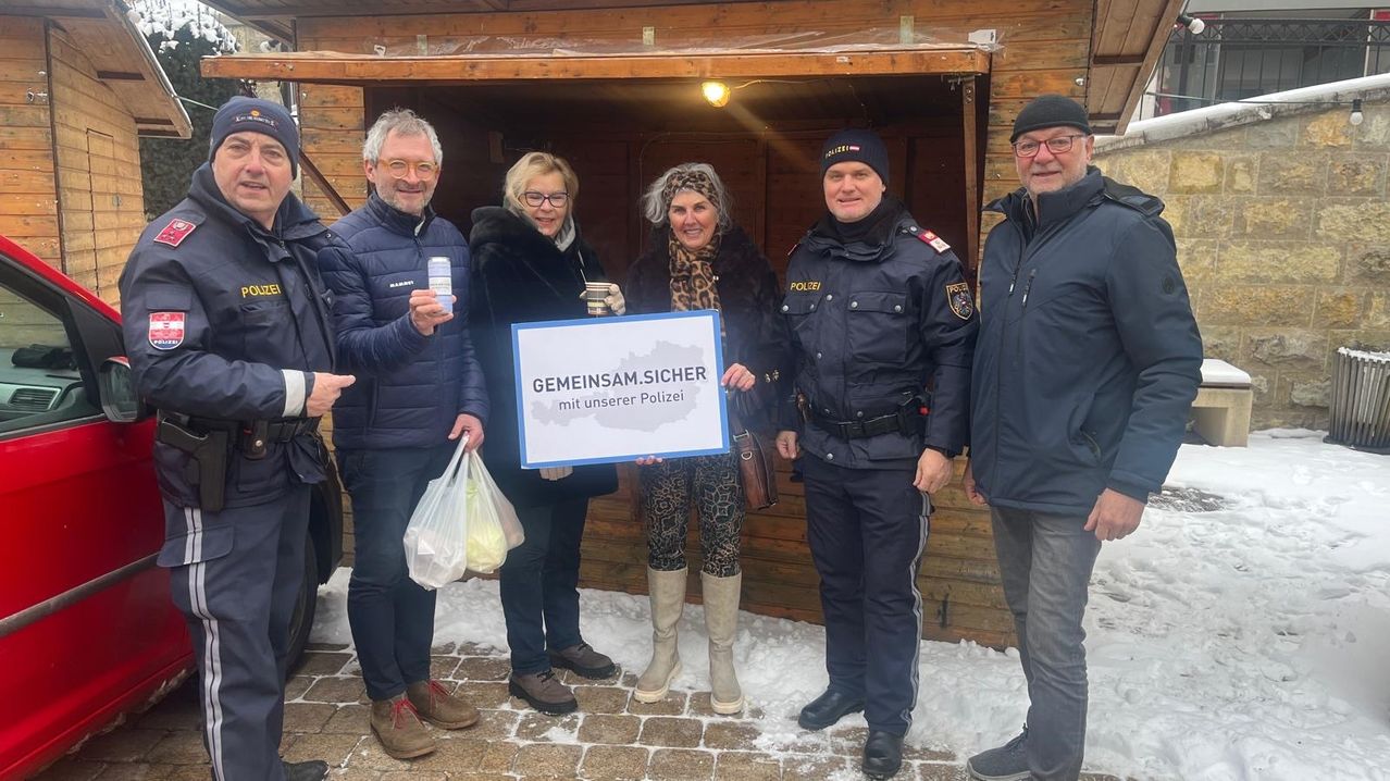 Eine Gruppe von Menschen steht in einem verschneiten Bereich und hält ein Schild mit der Aufschrift 'GEMEINSAM.SICHER mit unserer Polizei'. Sie sind alle in Winterkleidung gekleidet, und einer hält eine Plastiktüte.
