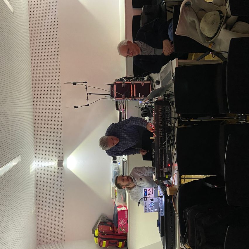 Drei Männer arbeiten an einem Tisch mit elektronischer Ausrüstung und einem Laptop. Einer sitzt, während die anderen stehen. Ausrüstung steht auf dem Tisch und eine rote Box ist auf dem Boden.