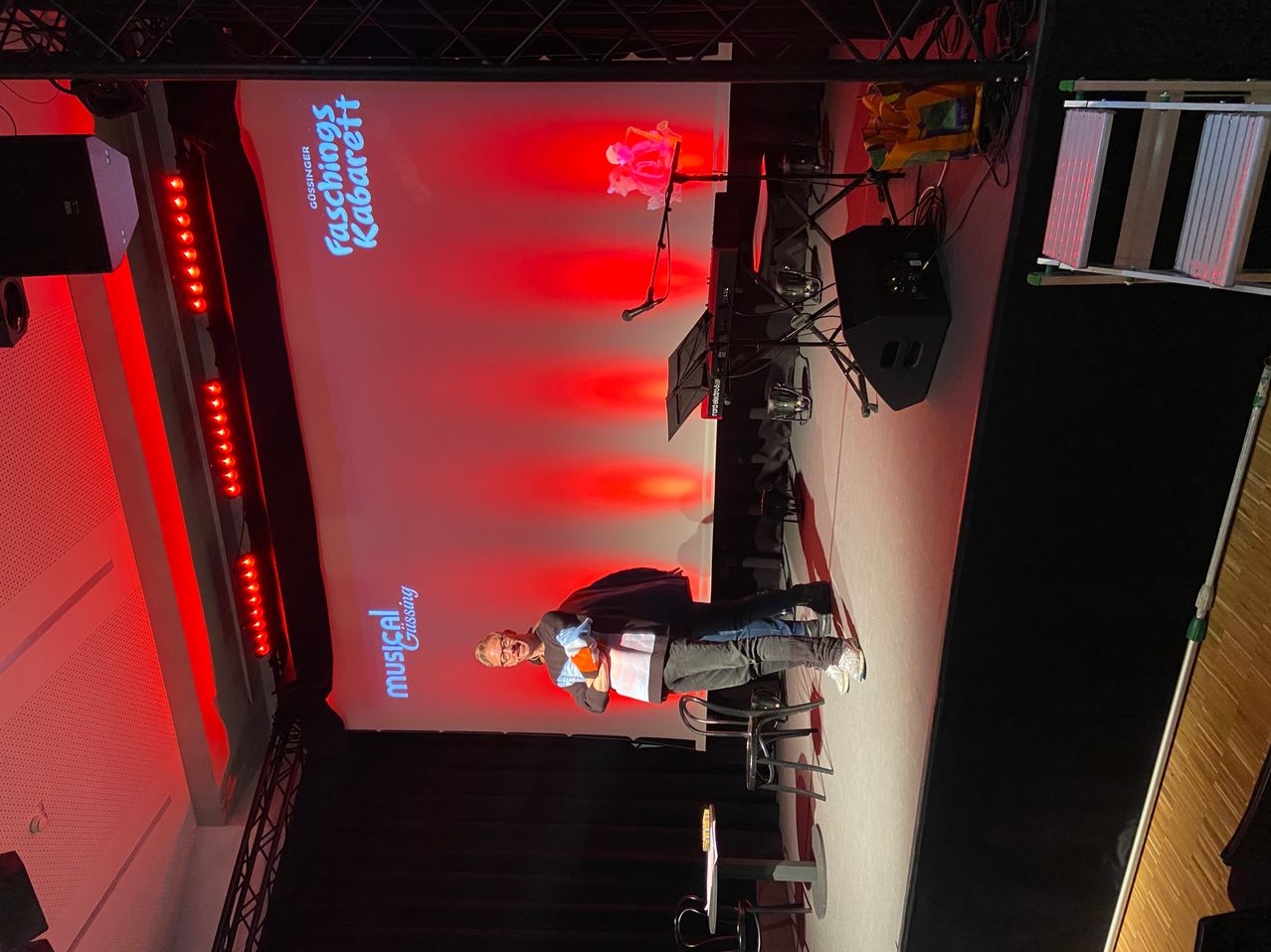 Eine Bühneneinrichtung mit einer Person auf einem Stuhl sitzend. Eine große Leinwand zeigt 'Fashionings Kabarett' und 'musical Crossing'. Rote Beleuchtung und verschiedene Musikgeräte sind vorhanden.