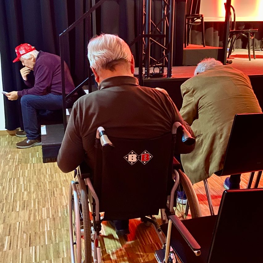 Drei Männer in einem Konferenzraum; einer im Rollstuhl, einer auf der Bühne sitzend und einer im Stuhl. Der Mann im Rollstuhl hat ein Logo auf dem Rücken.