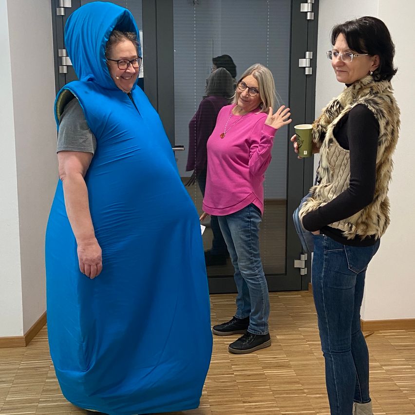 Drei Frauen stehen in einem Raum. Eine trägt eine blaue Kapuzenjacke, eine andere ein rosa Shirt und die dritte eine Pelzweste. Sie scheinen zu sprechen und lächeln.