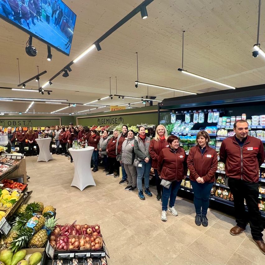 Eine Gruppe von Menschen steht in einem Geschäft, einige tragen burgunderrote Jacken, in der Nähe eines Regals voller Gemüse. Ein Fernseher ist an der Decke montiert.
