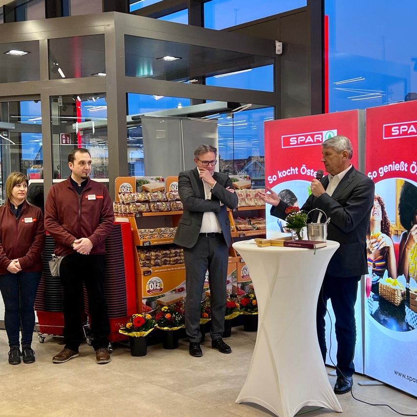 Ein Mann spricht in ein Mikrofon an einem Podium in einem Geschäft, mit drei Personen hinter ihm und einer Lebensmittelausstellung im Hintergrund.