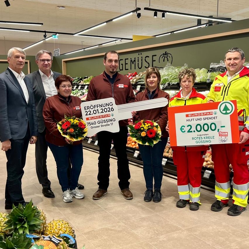 Eine Gruppe von Menschen steht in einem Geschäft und hält Blumen und einen großen Schlüssel. Der Schlüssel hat ein Schild mit der Aufschrift 'Eröffnung' und dem Datum '22.01.2026'. Zwei Frauen halten Schilder mit Blumen und eine sagt 'Eurospar'. Das Geschäft hat Obst und Gemüse und Beleuchtung.