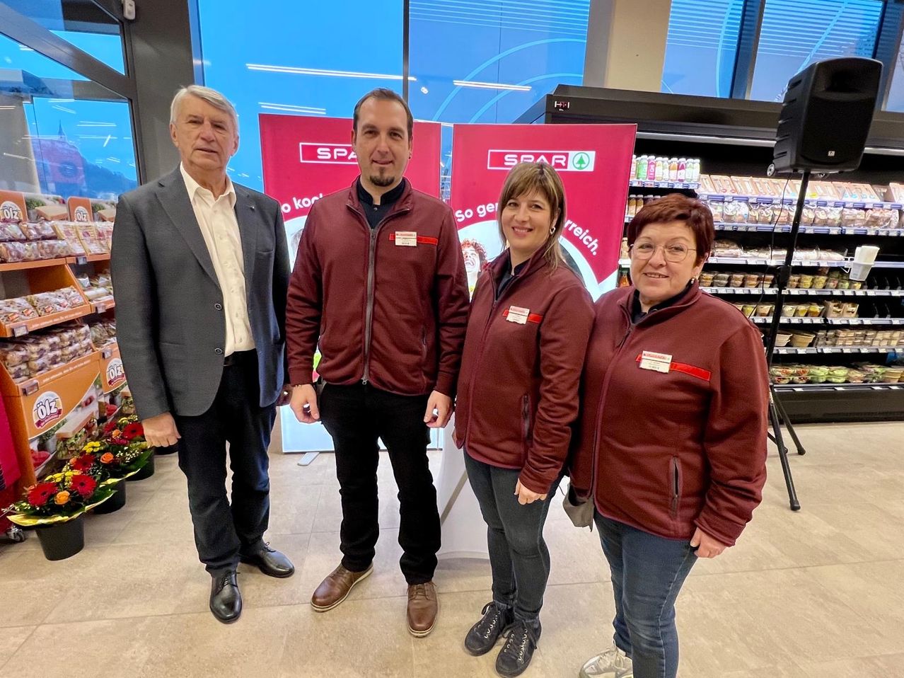 Vier Personen stehen in einem Geschäft mit einer lächelnden Frau, zwei Männern in Anzügen und einem Mann in einem Weinrot-Jacke. Hinter ihnen sind Banner und Regale mit Lebensmitteln.