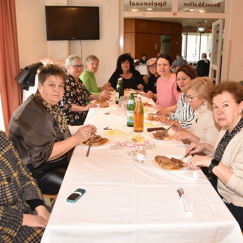 Eine Gruppe von Frauen in einem Esszimmer genießt eine Mahlzeit mit Tellern, Getränken und einem Fernseher im Hintergrund.