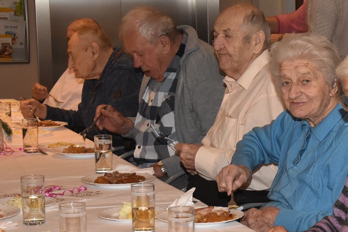 Eine Gruppe älterer Personen sitzt an einem Tisch und isst ihre Mahlzeiten. Jede Person hat einen Teller mit Essen, und es stehen Gläser mit Wasser auf dem Tisch.