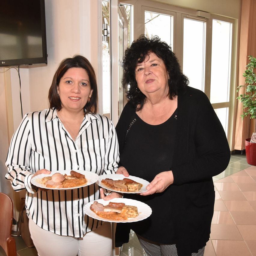 Zwei Frauen stehen nebeneinander und halten Teller mit Essen. Eine trägt ein gestreiftes Hemd und weiße Hose, die andere ein schwarzes Oberteil. Sie sind drinnen in der Nähe einer Glastür.