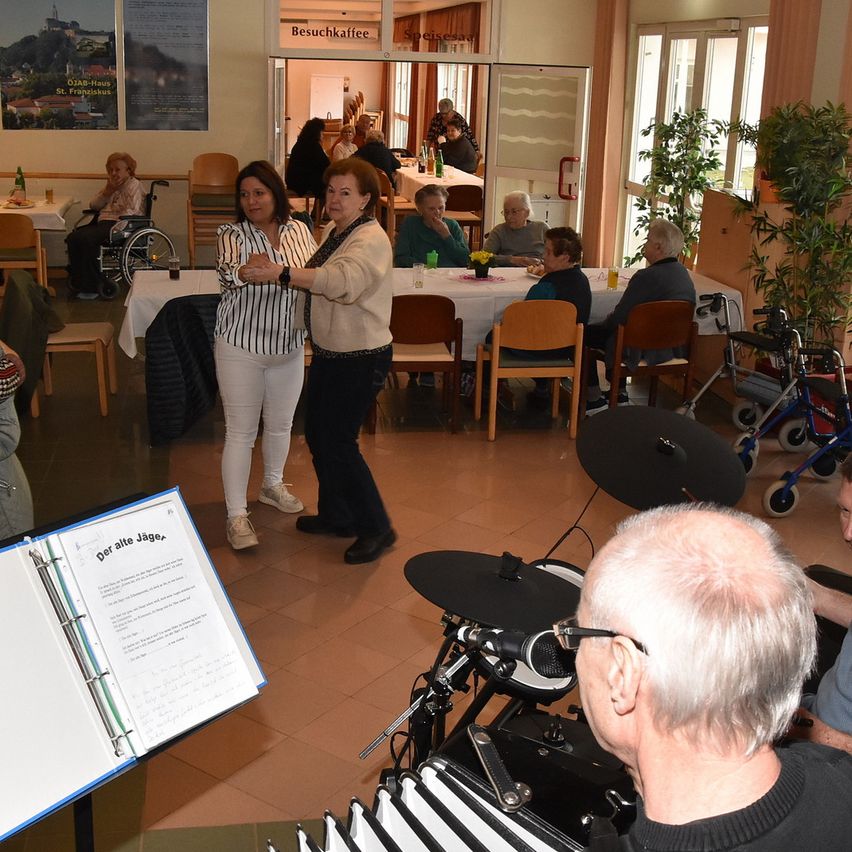Eine Gruppe älterer Menschen befindet sich in einem Raum mit Tischen, Stühlen und Musikinstrumenten. Zwei Frauen tanzen in der Mitte, während andere sitzen und zusehen. An der Wand hängt ein Plakat.