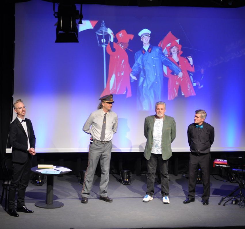 Fünf Männer stehen auf einer Bühne mit einer großen Leinwand im Hintergrund. Vier sind in Anzügen und Krawatten gekleidet. Die Leinwand zeigt drei Personen in roten Kostümen.