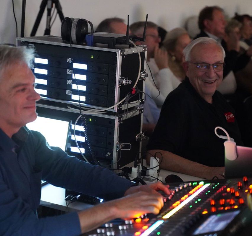 Zwei Männer arbeiten an einer Soundboard mit beleuchteten Bedienelementen, ein Geräteregal dahinter. Ein Publikum ist anwesend und beobachtet sie.