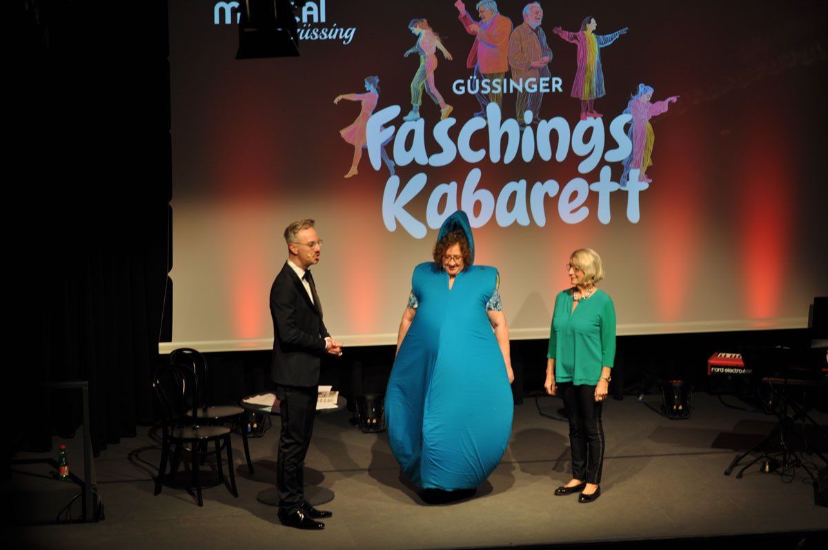 Drei Personen stehen mit einem großen blauen aufblasbaren Objekt auf der Bühne. Dahinter ist eine Leinwand mit der Aufschrift 'Faschings Kabarett' und einer farbenfrohen Zeichnung.