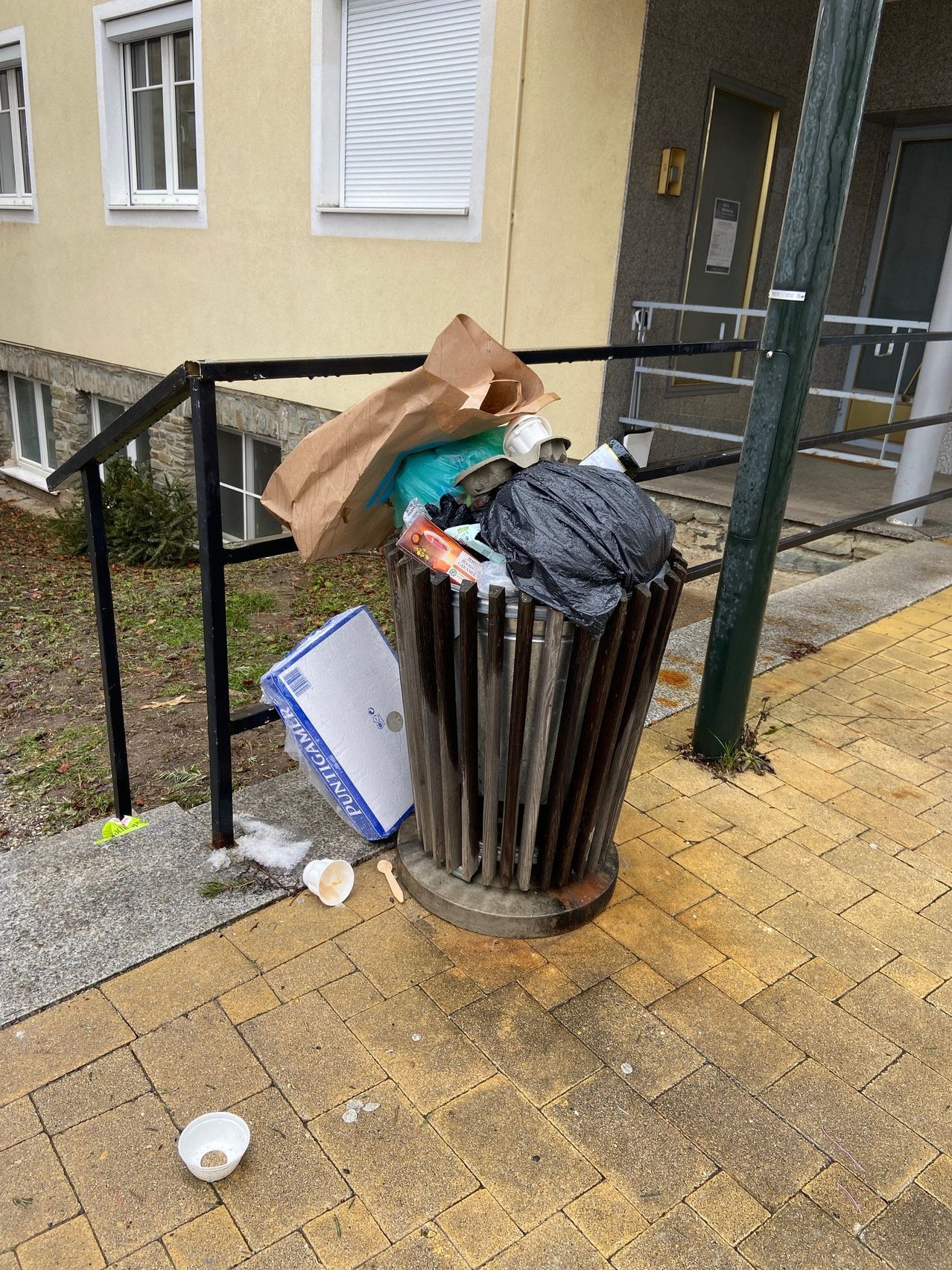 Ein überfüllter Mülleimer mit verschiedenen Abfällen, einschließlich eines Kartons, eines Plastiksacks und eines Papiersacks, vor einem Gebäude mit Treppen und Geländern.