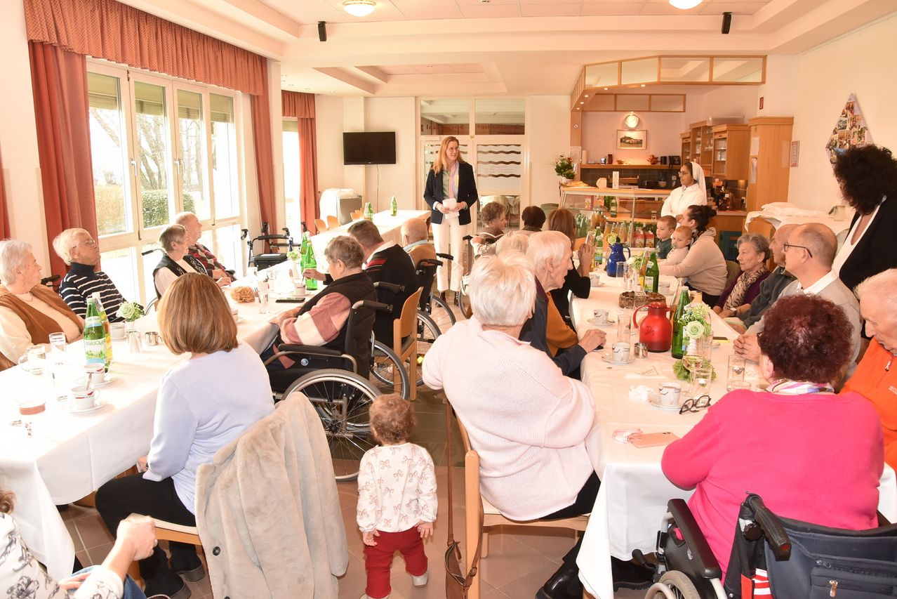 Eine Frau spricht an einem Mikrofon an einem langen Tisch in einem Speisesaal, um sie herum sitzen ältere Menschen, einige in Rollstühlen. Ein kleines Kind steht in der Nähe.