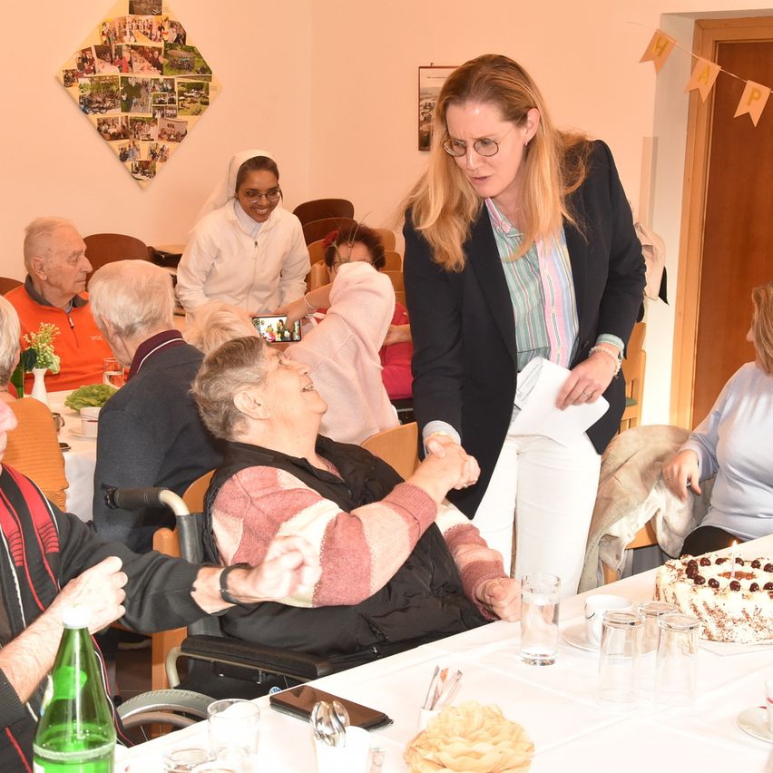 Eine Gruppe älterer Menschen ist zu einer Feier versammelt. Eine Frau im Rollstuhl lächelt und hält die Hand einer anderen Person. Eine Frau mit Brille liest von einem Papier. Ein Kuchen mit Kirschen steht auf dem Tisch.
