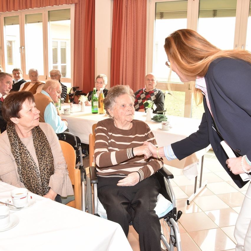 Eine ältere Frau im Rollstuhl begrüßt eine andere Frau. Sie sind in einem Essbereich mit mehreren älteren Menschen. Eine Frau steht neben dem Rollstuhl. Der Raum hat große Fenster und Tische mit Blumen und Tassen.