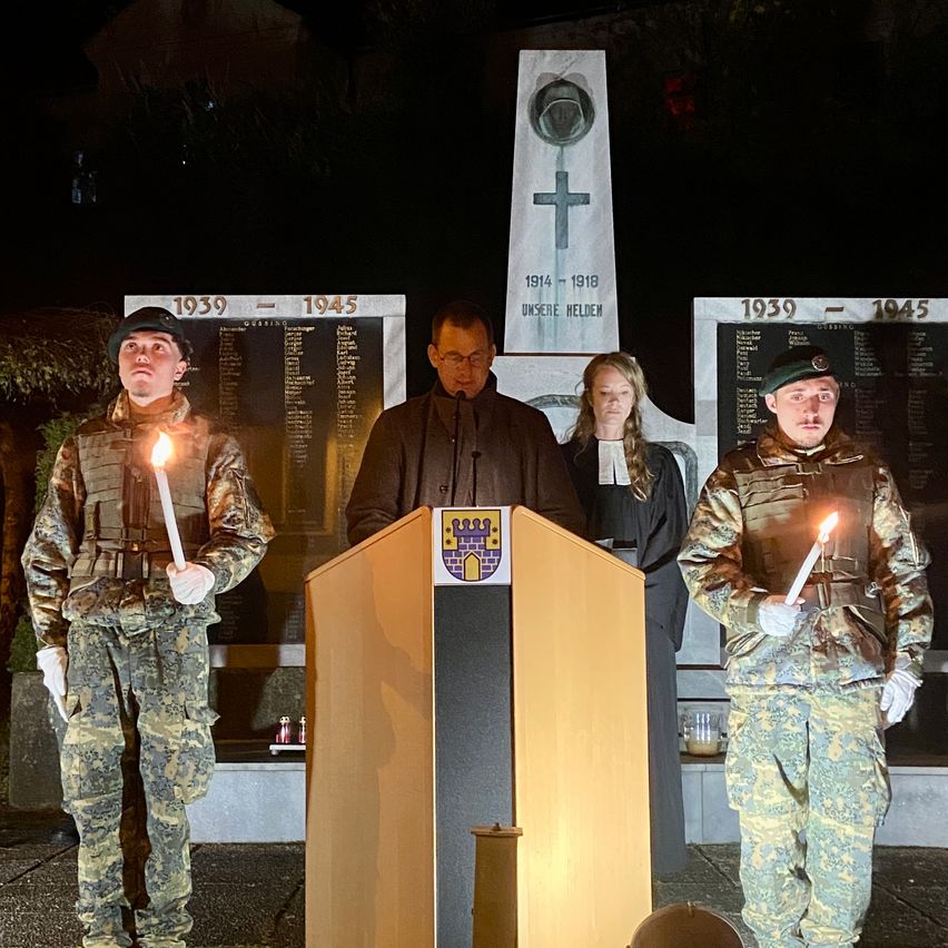 Three individuals in military uniforms stand at a podium at night, holding lit candles. Behind them is a memorial with a cross and the dates 1939-1945 on it. A man and a woman stand to the sides of the podium.