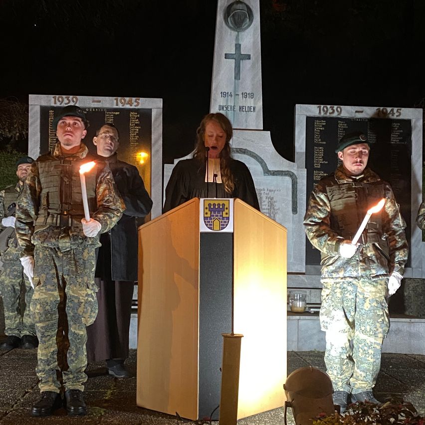 At night, a woman speaks at a podium in front of a memorial. Two soldiers stand behind her, holding lit candles. The memorial has a cross and two plaques with dates.