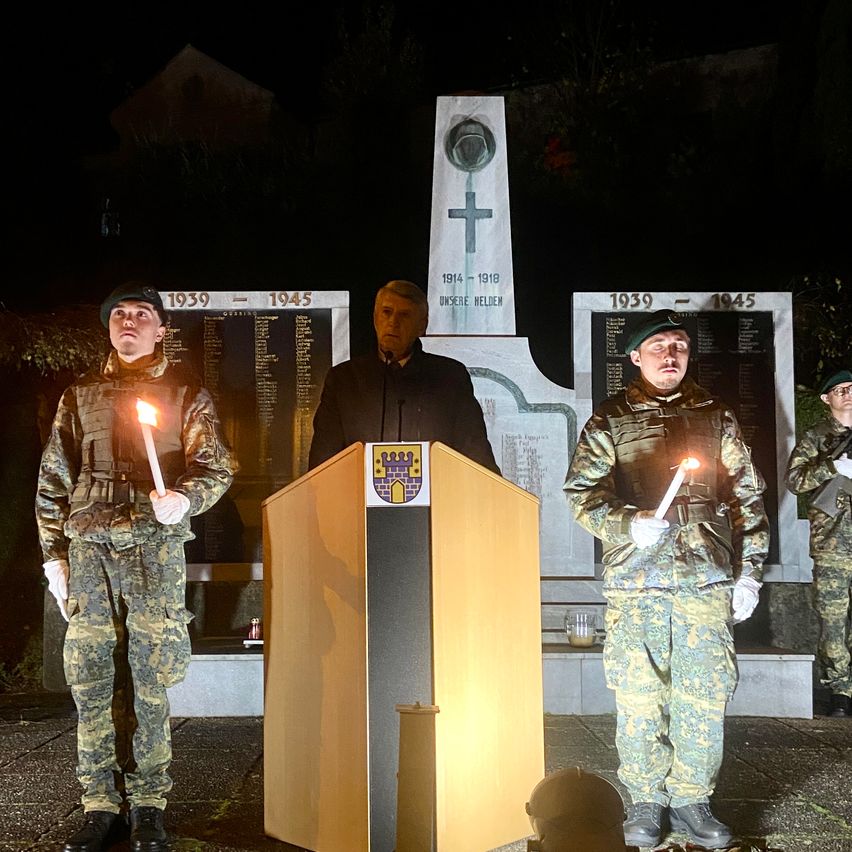 Four individuals in military uniforms stand at a memorial. Two hold lit candles. A man speaks at a podium. A cross and plaques with years 1914-1918 and 1939-1945 are visible.