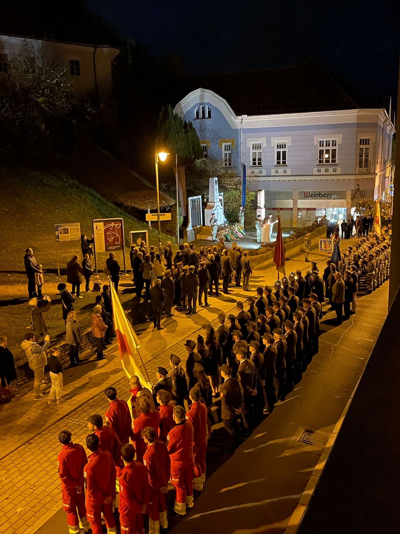 A nighttime gathering of people in red uniforms is occurring in front of a building. Some individuals are holding flags, and others are standing on the side. A statue is located in front of the building.