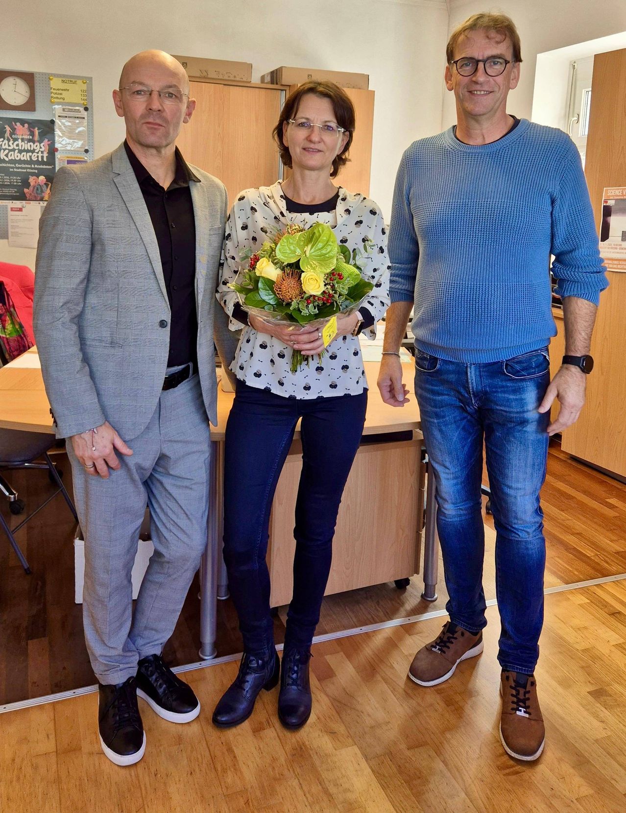 Three people stand next to each other in a room. The woman in the middle holds a bouquet of flowers. The man on the left wears a suit and leather shoes. The man on the right wears a sweater and jeans.