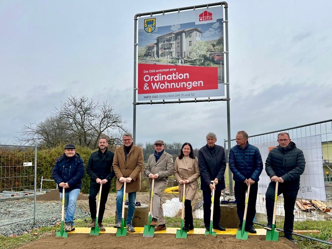 Eine Gruppe von Menschen in formeller Kleidung steht auf einer Baustelle und hält Schaufeln. Ein Banner mit einem Gebäudebild ist auf einer Metallstruktur montiert. Das Banner lautet 'Die OSG errichtet eine Ordination & Wohnungen'.