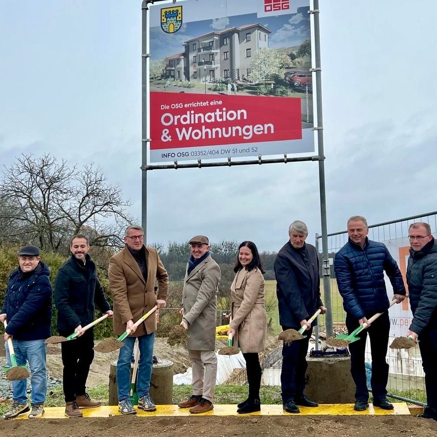 Eine Gruppe von Menschen in Mänteln steht auf einem Erdfleck, hält Schaufeln und posiert für ein Foto vor einem großen Schild für ein Wohnprojekt.