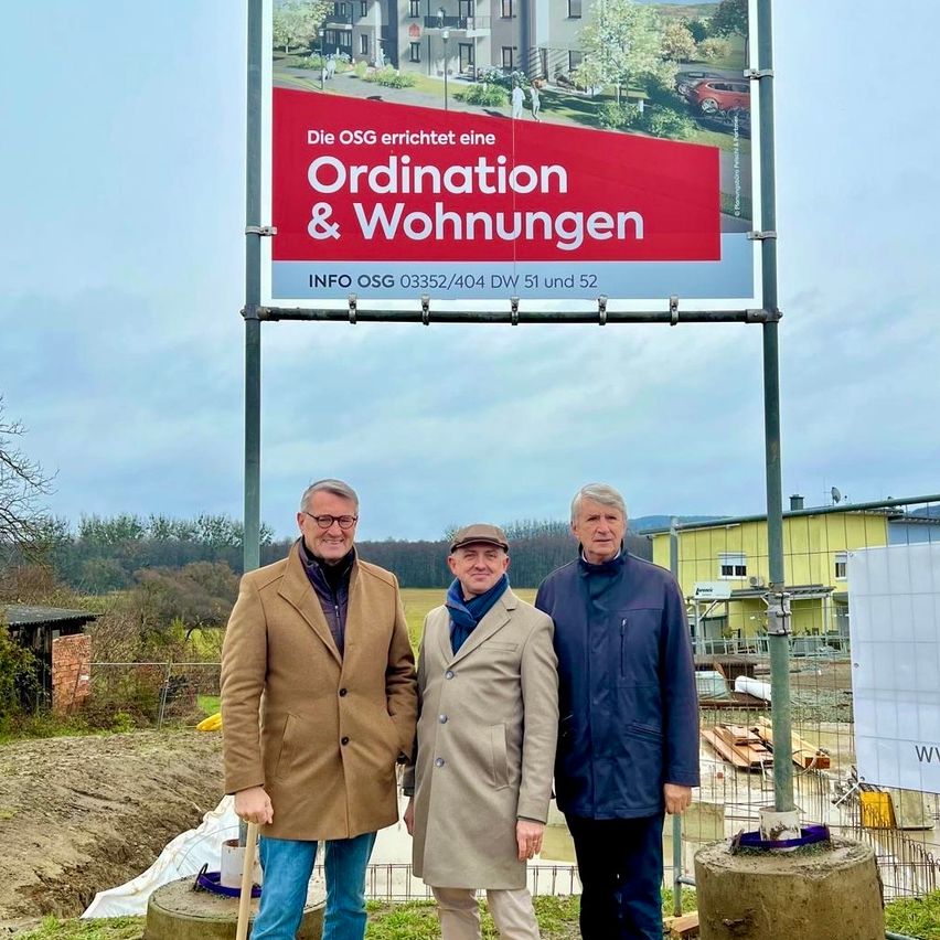 Drei Männer stehen vor einer Baustelle. Hinter ihnen steht ein Schild, das ein Wohnprojekt von OSG anzeigt. Ein Hausmodell ist zu sehen, und das Schild trägt die Aufschrift 'Ordination & Wohnungen' mit Kontaktinformationen.
