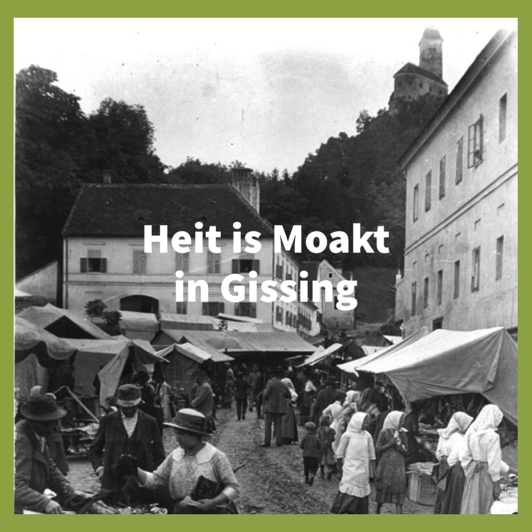 Black and white photo of a bustling marketplace in Gising. People are shopping at stalls, some wearing traditional clothing. In the background, buildings and a castle are visible.